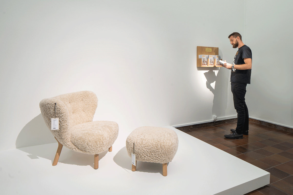 Beige armchair and ottoman on a white platform with a person holding a framed picture in the background.