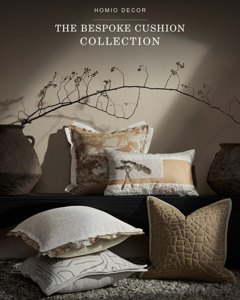 A styled collection of decorative throw pillows in warm neutral tones, arranged on a dark bench against a beige wall with dried branches and ceramic vases. 