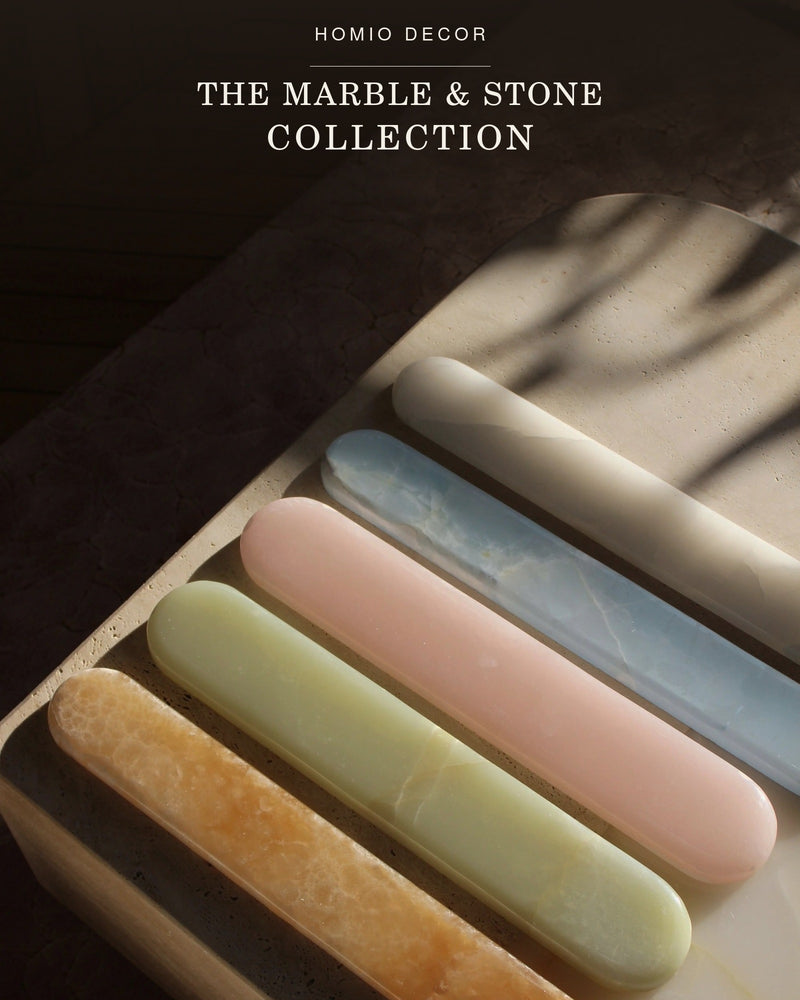 Five elongated stone or marble bar samples in cream, pale blue, blush pink, pale yellow, and warm tan are arranged diagonally on a textured surface under soft shadows. 