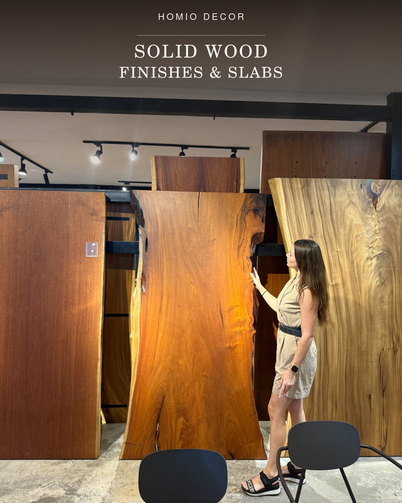 A woman stands beside large solid wood slabs in a showroom, pointing at a warm honey-brown live edge wood panel. 