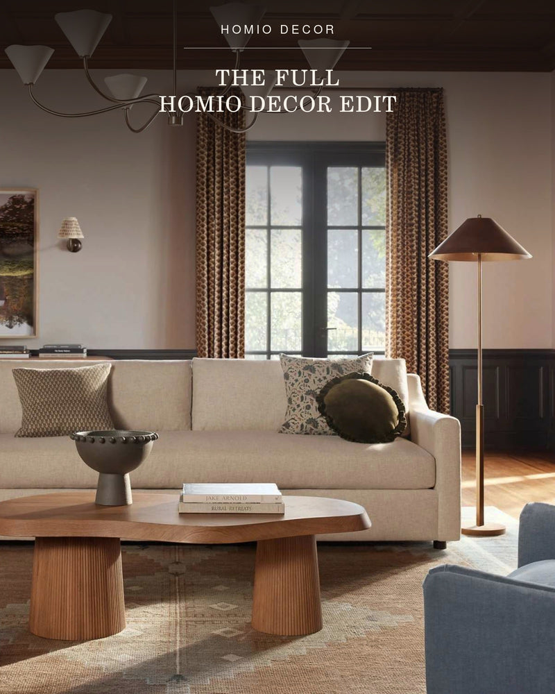 A styled interior scene showing a cream sofa in a living room with patterned pillows, a wooden coffee table, curtains, a floor lamp.