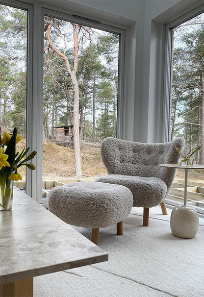 Gray upholstered chair and ottoman in a room with large windows overlooking a forest.