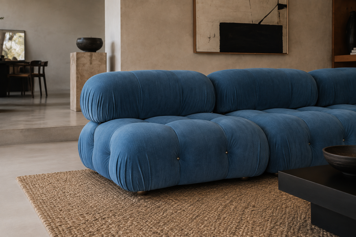 A modern tufted modular sofa in a rich blue velvet fabric, styled in a minimalist, warm-toned living room with a woven jute rug and concrete floors.