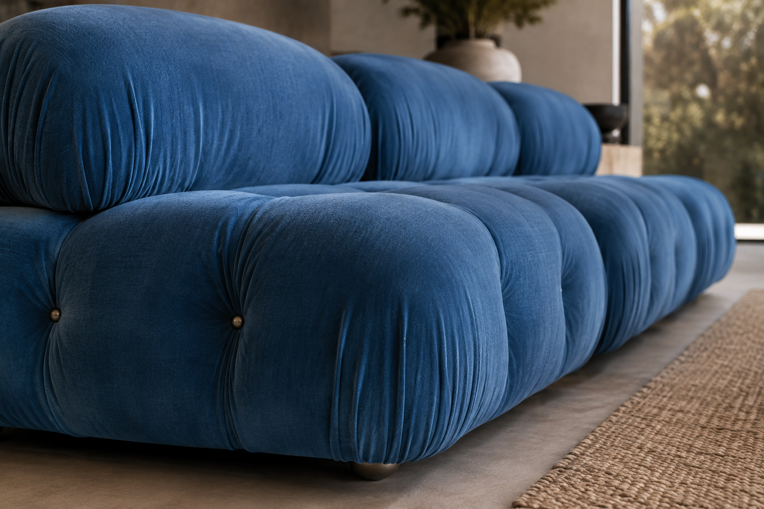 A close-up of a modern tufted sofa in rich blue velvet, featuring rounded segmented cushions and small gold button accents, set in a contemporary living room with a woven rug and window view.